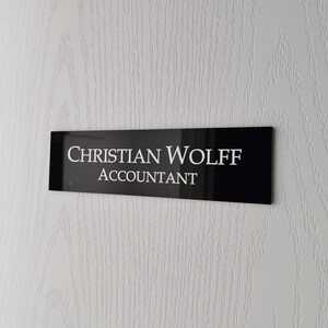May include: A rectangular black sign with a glossy finish featuring the white text Christian Wolff Accountant in a serif font. The sign is mounted horizontally on a white surface with a subtle vertical wood grain texture.
