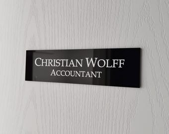 May include: A rectangular black sign with a glossy finish featuring the white text Christian Wolff Accountant in a serif font. The sign is mounted horizontally on a white surface with a subtle vertical wood grain texture.