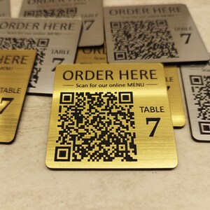 May include: Gold and silver metal table signs with the text "ORDER HERE" and a QR code. Each sign also displays "TABLE 7". The signs are designed to be scanned for an online menu.