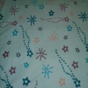 May include: White fabric with a repeating pattern of blue, pink, and gray stars and fireworks. The stars are of various sizes and the fireworks are stylized.