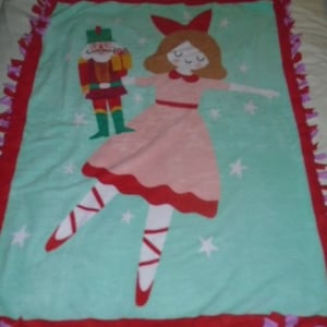 May include: A turquoise blue fleece blanket with a red, white, and pink ballerina and a nutcracker. The blanket has a red border with purple fringe.