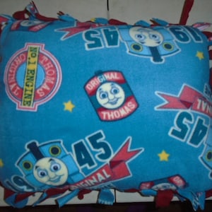 May include: A square, blue fleece pillow featuring images of Thomas the Tank Engine. The pillow has red and blue knotted edges. The design includes the words "Original Thomas" and "No. 1 Engine".