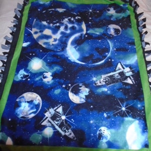 May include: A blue fleece tie blanket with a space theme. The blanket features planets, stars, and spaceships on a dark blue background. The blanket is trimmed with green and black fringe.