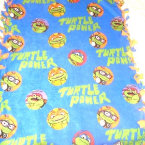May include: A blue fleece tie blanket with a Teenage Mutant Ninja Turtles theme. The blanket features a repeating pattern of the Teenage Mutant Ninja Turtles characters in green, orange, and black, with the words "Turtle Power" repeated throughout.