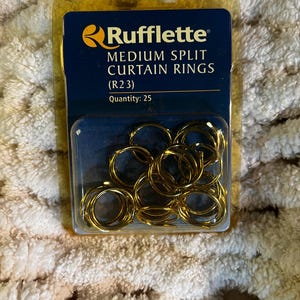 May include: A package of 25 gold-colored Rufflette medium split curtain rings, model R23. The product is contained in a clear plastic blister pack with a dark blue cardboard backing featuring the brand name and product details in white text.