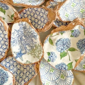 May include: A collection of decorated oyster shells. The shells feature various blue and white patterns, including floral and geometric designs. Some shells have gold-colored edges. The designs include hydrangeas, starfish, and coral.