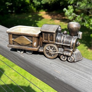May include: A bronze-colored ceramic train planter featuring a locomotive engine attached to a rectangular cargo car. The train has detailed wheels, a smokestack, and a hollow interior compartment for plants, finished with a metallic patina.