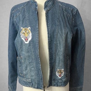 Peut inclure: Veste en jean bleu moyen avec un col montant et une fermeture éclair sur toute la longueur. La veste comporte deux écussons de tête de tigre, un de chaque côté de la poitrine. Les écussons ont une bordure blanche et sont dans les tons de jaune, orange et noir.