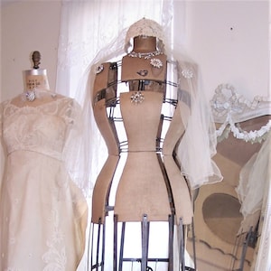 May include: A vintage dress form with a metal cage skirt and a white lace veil. The dress form is standing on a wooden floor in front of a white mirror.