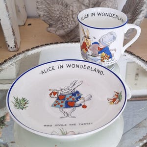 May include: A white teacup and saucer set with blue trim and illustrations from Alice in Wonderland. The teacup features the Mad Hatter and the March Hare, while the saucer features the White Rabbit holding a scroll and a playing card. The text on the teacup reads "Alice in Wonderland" and the text on the saucer reads "Alice in Wonderland. Who stole the tarts?"