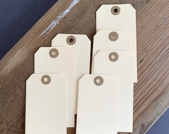 Puede incluir: Un conjunto de seis etiquetas rectangulares de papel color crema con las esquinas superiores recortadas. Cada etiqueta presenta un orificio circular en la parte superior reforzado con un ojal de papel marrón. Las etiquetas están dispuestas de forma escalonada.
