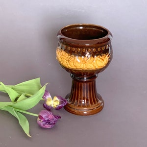 May include: A brown ceramic planter with a pedestal base. The planter has a raised design of yellow flowers around the top edge.