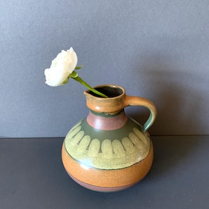 Könnte beinhalten: Ein kleiner, runder Keramikkrug mit Henkel, mit einer weißen Blume. Der Krug hat ein braunes, grünes und orangefarbenes Farbschema mit einem dekorativen Muster in der Mitte. Die Blume ist weiß mit einem grünen Stiel.