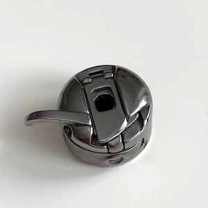 May include: A close-up of a dark gray metal sewing machine bobbin case. The case has a curved handle and a hinged top. The bobbin case is designed to hold the bobbin thread for a sewing machine.