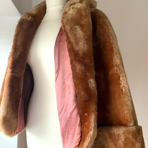 Puede incluir: Un abrigo vintage de piel sint&eacute;tica marr&oacute;n, exhibido en un maniqu&iacute; blanco. El abrigo tiene un cuello ancho, mangas largas con pu&ntilde;os doblados y un forro de sat&eacute;n rosa. El color c&aacute;lido y neutro del abrigo es perfecto para el clima fresco.