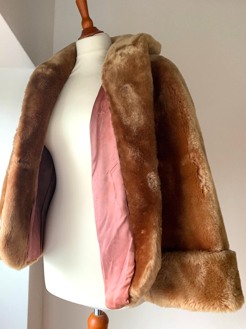 Puede incluir: Un abrigo vintage de piel sint&eacute;tica marr&oacute;n, exhibido en un maniqu&iacute; blanco. El abrigo tiene un cuello ancho, mangas largas con pu&ntilde;os doblados y un forro de sat&eacute;n rosa. El color c&aacute;lido y neutro del abrigo es perfecto para el clima fresco.