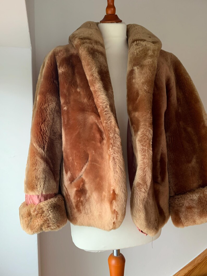 Puede incluir: Un abrigo vintage de piel sint&eacute;tica marr&oacute;n con cuello ancho y pu&ntilde;os. El abrigo se exhibe en un maniqu&iacute;, mostrando su textura afelpada y color c&aacute;lido. Los pu&ntilde;os revelan un forro rosa, agregando un sutil contraste.