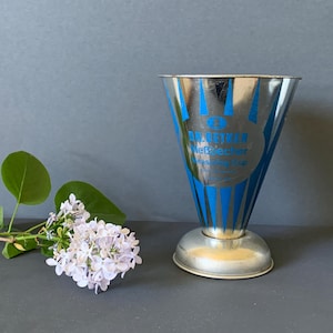 Könnte beinhalten: Ein Vintage-Messbecher aus Metall mit blauen Streifen und Text. Der Becher hat eine breite Oberseite und einen kleinen Sockel. Ein Zweig Fliederblüten mit grünen Blättern liegt neben dem Becher.