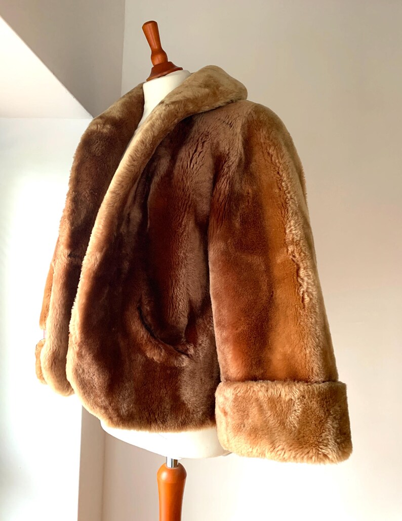 Puede incluir: Chaqueta de piel sint&eacute;tica marr&oacute;n con cuello ancho y pu&ntilde;os doblados. La chaqueta se exhibe en un maniqu&iacute; blanco y tiene un aspecto suave y texturizado. La chaqueta tiene un solo bolsillo.