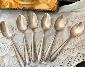 May include: Six silver spoons with ornate handles are arranged on a white lace tablecloth. The spoons are in a box lined with yellow satin.
