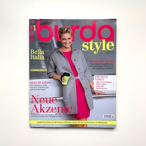 Könnte beinhalten: Ein Magazincover mit einer Frau, die ein rosa Kleid und einen grauen Mantel trägt. Das Magazin heißt "Burda Style" und hat den Text "Bella Italia" und "Neue Akzente" auf dem Cover.