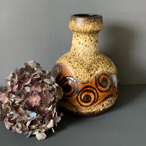 Könnte beinhalten: Eine Vintage-Keramikvase mit einer strukturierten, gesprenkelten Oberfläche in Braun- und Beigetönen. Die Vase hat ein dekoratives Muster aus braunen und orangefarbenen Wirbeln. Eine getrocknete Hortensienblüte in Lila- und Brauntönen liegt neben der Vase.