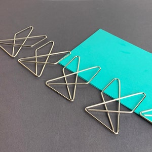 Pode incluir: Cinco clipes de papel prateados em forma de triângulo com um design de cruz. Os clipes estão dispostos em uma fila sobre uma superfície cinza escura com um pedaço de papel azul turquesa atrás.