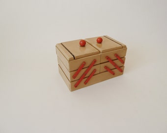 Puede incluir: Una caja de madera con cuatro compartimentos, cada uno con una perilla roja. La caja tiene un acabado beige y costuras rojas en forma de cruz en la parte delantera.