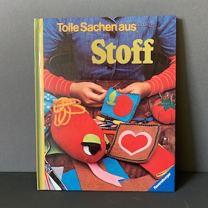 Könnte beinhalten: Buchcover mit dem Titel "Tolle Sachen aus Stoff" in gelben Buchstaben. Das Cover zeigt ein Jeanshemd und rotärmelige Arme, mit verschiedenen Filzarbeiten wie einer roten Schlange und einem blauen Quadrat.
