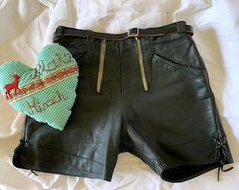 Puede incluir: Pantalones cortos de cuero negro con dos cremalleras verticales en la parte delantera, un cinturón fino de cuero negro y detalles de cordones laterales. Junto a los pantalones hay un cojín en forma de corazón de cuadros verdes y blancos bordado con un ciervo rojo y el texto Platz Hirsch.