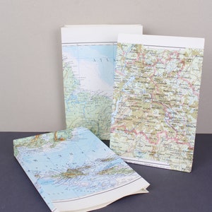 Puede incluir: Se muestran tres bolsas de papel cubiertas con dise&ntilde;os de mapas. Los mapas muestran detalles geogr&aacute;ficos en varios colores. Una bolsa est&aacute; abierta, revelando el interior del mapa. Las bolsas tienen la parte superior blanca.