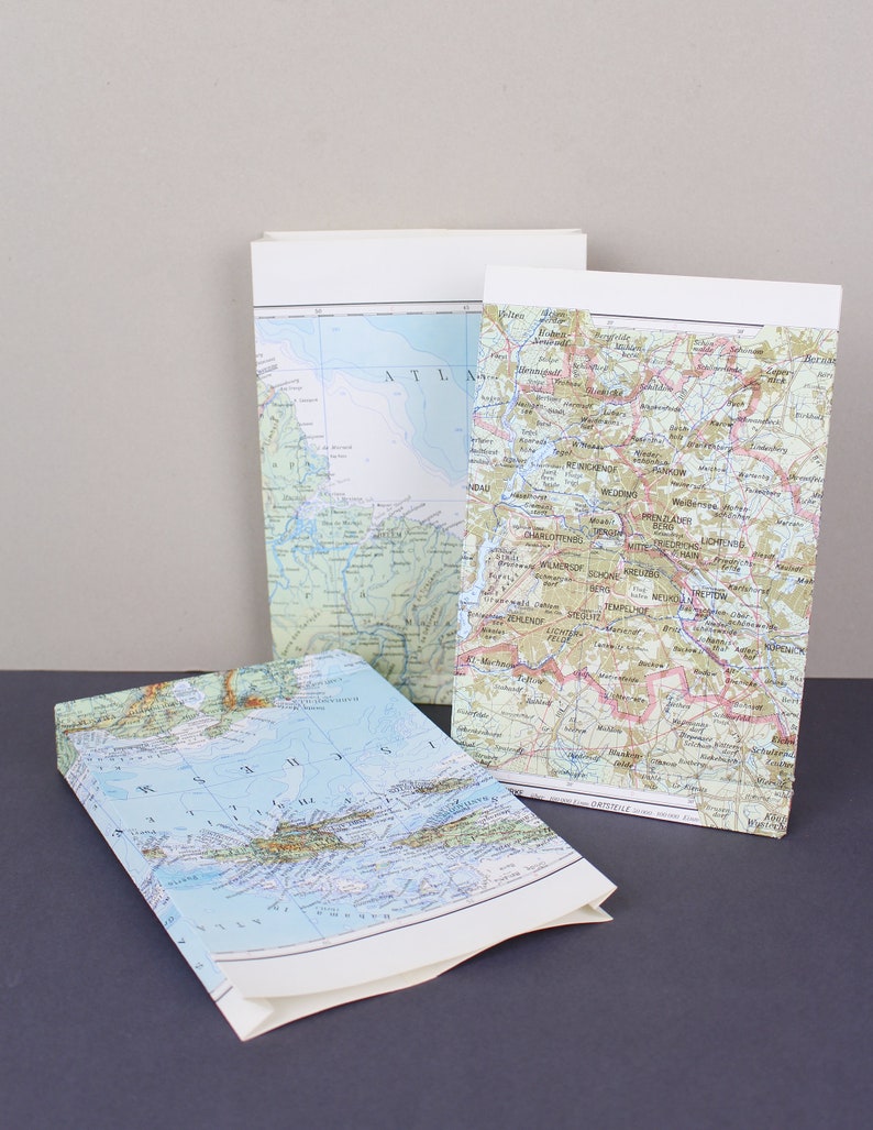 Puede incluir: Se muestran tres bolsas de papel cubiertas con dise&ntilde;os de mapas. Los mapas muestran detalles geogr&aacute;ficos en varios colores. Una bolsa est&aacute; abierta, revelando el interior del mapa. Las bolsas tienen la parte superior blanca.