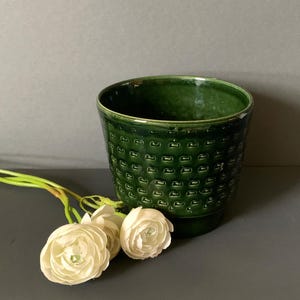 Puede incluir: Una maceta de cerámica verde oscuro con un patrón geométrico texturizado. La maceta es cilíndrica con un borde ligeramente ensanchado. Flores blancas con tallos verdes se colocan cerca de la maceta.
