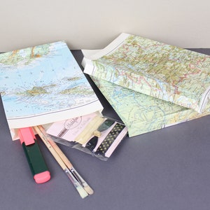 Puede incluir: Tres cajas de regalo cubiertas con mapas, dos apiladas y una abierta, que revelan materiales de arte. La caja abierta contiene un resaltador rosa, pinceles y una peque&ntilde;a bolsa con cremallera y cinta decorativa. Sobre una superficie gris.