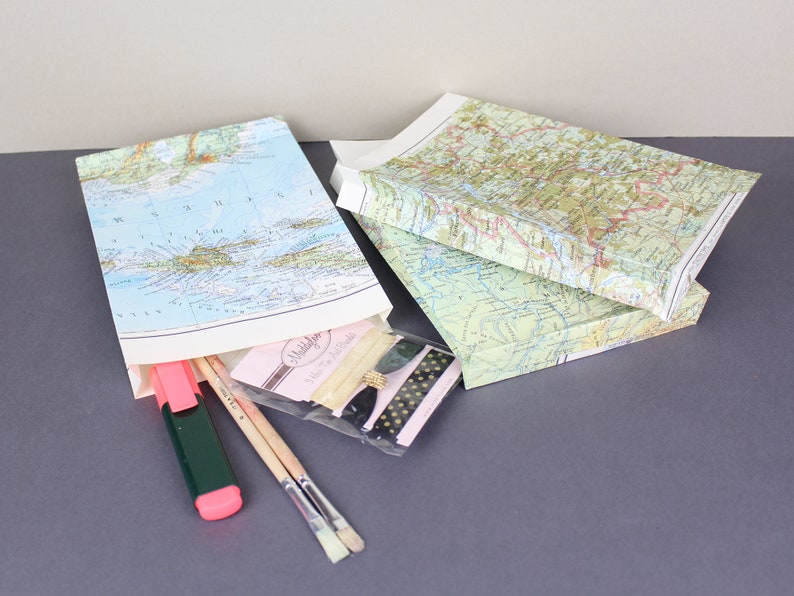 Puede incluir: Tres cajas de regalo cubiertas con mapas, dos apiladas y una abierta, que revelan materiales de arte. La caja abierta contiene un resaltador rosa, pinceles y una peque&ntilde;a bolsa con cremallera y cinta decorativa. Sobre una superficie gris.