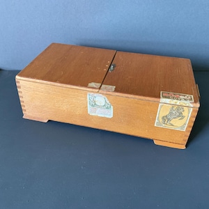 Könnte beinhalten: Eine rechteckige Holzkiste mit Scharnierdeckel und hellbraunem Finish. Die Kiste hat einen kleinen Metallverschluss und ist mit mehreren Vintage-Etiketten verziert, darunter eines mit einer Pferdeillustration. Die Kiste steht auf einer blauen Oberfläche.