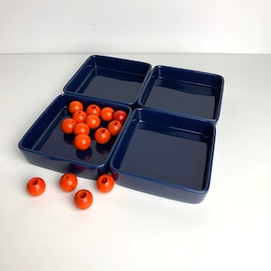 May include: Four square blue ceramic dishes with a pile of orange round beads in one of the dishes.