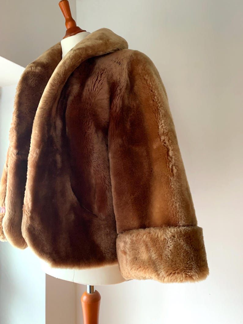 Puede incluir: Un abrigo de piel sint&eacute;tica marr&oacute;n con un cuello ancho y pu&ntilde;os doblados. El abrigo se exhibe en un maniqu&iacute;, mostrando su textura afelpada y su color c&aacute;lido. El dise&ntilde;o del abrigo presenta una silueta cl&aacute;sica.