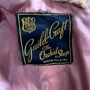 Puede incluir: Primer plano de una etiqueta de "Guild-Craft The Orchid Shop" en Wichita Falls, Texas. La etiqueta es negra con letras doradas y un emblema dorado. El forro es rosa con un patr&oacute;n de lazo.