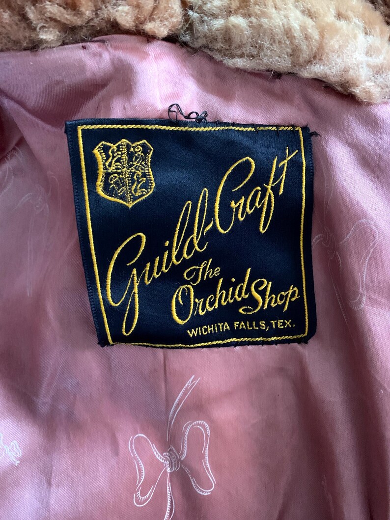 Puede incluir: Primer plano de una etiqueta de "Guild-Craft The Orchid Shop" en Wichita Falls, Texas. La etiqueta es negra con letras doradas y un emblema dorado. El forro es rosa con un patr&oacute;n de lazo.