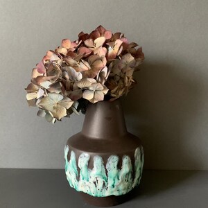 Könnte beinhalten: Eine braune Keramikvase mit türkisfarbenem und weißem Tropfglasur-Design, die getrocknete Hortensien mit tan- und lilafarbenen Blütenblättern enthält. Die Vase hat einen abgerundeten Körper und einen schmalen Hals, wodurch ein dekoratives Blumenarrangement entsteht.