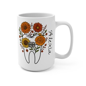 Puede incluir: Taza de cerámica blanca con un diseño de diente con brackets, rodeada de flores naranjas y amarillas y hojas verdes. El nombre "Alexia" está escrito en escritura negra en el lateral.