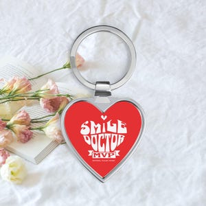 Puede incluir: Llavero en forma de corazón con centro rojo y borde metálico plateado. El centro rojo presenta las palabras "SMILE DOCTOR MVP" en letras blancas estilizadas. El llavero se muestra sobre una superficie blanca con flores y un libro.