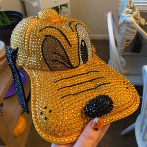 May include: A yellow baseball cap with a cartoon dog face design covered in rhinestones. The dog's eyes are black and white, and the nose is black.