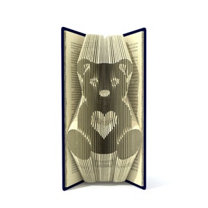 May include: A book with pages folded into the shape of a teddy bear holding a heart. The book is open and the pages are folded to create a 3D effect. The book is blue on the outside.
