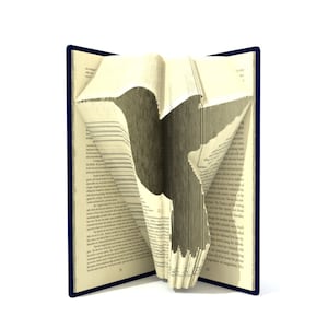 Puede incluir: Un libro con las páginas dobladas en forma de pájaro. El libro está abierto y las páginas están dobladas para crear una silueta de un pájaro. El libro está sobre un fondo blanco.