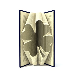 Könnte beinhalten: Ein Buch mit Seiten, die zu einem vierblättrigen Kleeblatt gefaltet sind. Das Buch ist geöffnet und das Kleeblatt ist in der Mitte des Buches sichtbar. Das Buch hat einen blauen Einband.