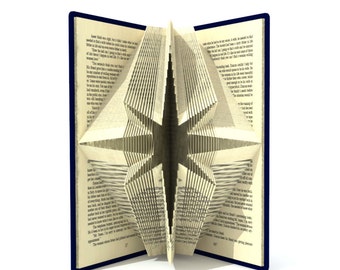 May include: A book with pages folded into a 3D star shape. The book is open and the star is visible in the center of the image. The book has a blue cover.