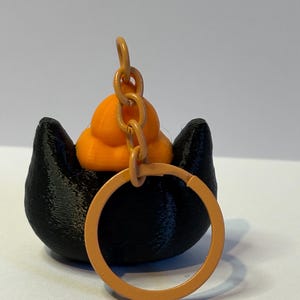 以下が含まれることがあります： 黒い猫の形をしたベースとオレンジ色のアクセントが特徴の3Dプリントキーホルダー。猫の頭には小さなオレンジ色のカボチャがあり、オレンジ色のキーリングが付いています。装飾的なアクセサリーとして設計されています。