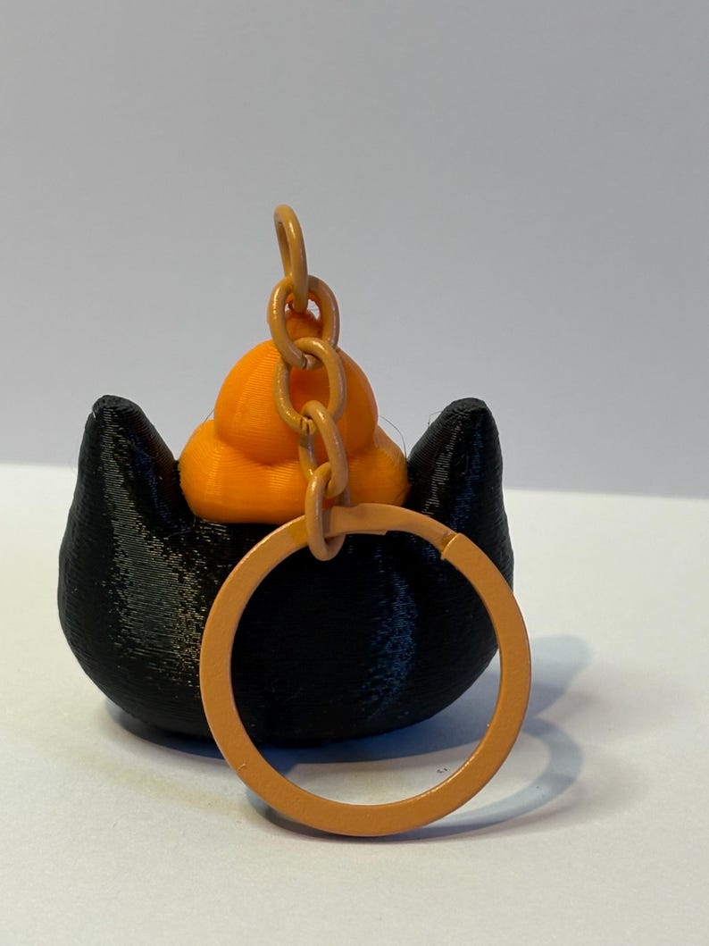 以下が含まれることがあります： 黒い猫の形をしたベースとオレンジ色のアクセントが特徴の3Dプリントキーホルダー。猫の頭には小さなオレンジ色のカボチャがあり、オレンジ色のキーリングが付いています。装飾的なアクセサリーとして設計されています。